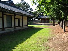 In a sunny day, a Korean traditional wooden building painted with white and dark red stands on a grass field. Luxuriant trees are seen on the right while a gate is shown at a distance.