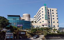 A large seven-story hospital complex on a slope that consists of about two buildings. The wall of one on the left is covered with blue glasses, and the other building with round corners is covered with beige bricks. Large green vertical signs are attached on the wall of the latter. The signs say