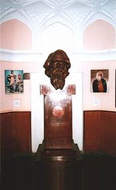 A bronze bust of a middle-aged and forward-gazing bearded man supported on a tall rectangular wooden pedestal above a larger plinth set amidst a small ornate octagonal museum room with pink walls and wooden paneling; flanking the bust on the wall behind are two paintings of Tagore: to the left, a costumed youth acting a drama scene; to the right, a portrait showing an aged man with a large white beard clad in black and red robes.