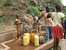 Fotografia que descreve um adulto e cinco crianças que enchem garrafas de água em uma bomba de água do metal rural com base de concreto, na parte inferior de uma encosta rochosa íngreme