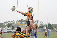 A female player in yellow and green kit and wearing a white scrum cap, jumps to collect a ball while supported by team mates.