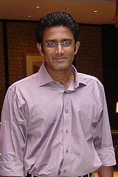 Brown skinned man with black, combed back hair, wearing rimless glasses and an open-necked lavender shirt with rolled up sleeves.