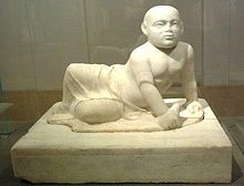 Beige marble statue of a stout young child aged about two years old lying on his left side. The child's head is shaved, his eyes gaze over the viewer's shoulder and his lower body is covered in a draping cloth that hangs limply between his flexed feet. The child supports his torso with his left hand in which he holds an unidentifiable object, he also holds a small bird in his right hand. The sculpture rests on a heavy socle inscribed with barely visible letters spanning the upper part of the socle vertically.