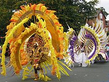 Part of a West Indian carnival procession passes along a public road in front of trees (to the left) and red-brick houses (to the right). In front is a participant wearing a gold helmet and vestigial armour. Above and around her is an enormous shield-shaped yellow, gold and red contraption supported by spokes and with many large feathers. Behind her is another participant in a white dress with a similar-shaped feathered shield contraption in yellow, brown and purple worn the other way up.
