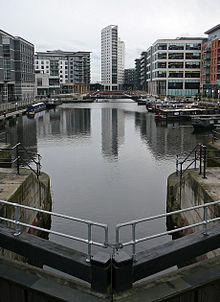 A night-time scene of a dock containing a number of moored canal-boats to left and right and railings around the edges. At the front is part of a lock gate and steps leading down to the water. Around most of the dock are multi-storey modern buildings, some with lighted ground-floors and seats and decorative objects outside. The most prominent of these, at the far end, is a twenty-storey building with curving façades.