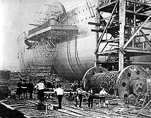 Uma fotografia velha que mostra um grande navio de pás de ferro a ser lançado de lado, com operários empurrando grandes baulks de madeira sob um grande tambor de correntes de ferro