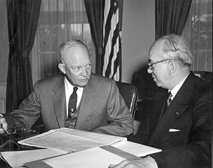 Two men in suits at a table covered in papers. There is an American flag in the background.