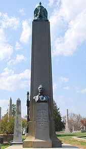 A large obelisk in a graveyard, with a bust of Tyler. The black structure visible behind the left side of Tyler's obelisk encloses the grave of James Monroe.