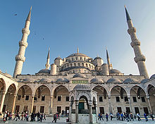 A building with four visible minarets, numerous domes, and vaulted arcades fronted by a courtyard with passer-by