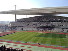 A stadium with a football pitch and track surrounded by multi-tier seating and covered by a large hanging roof