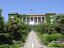 An ivy-covered neoclassical building atop a hill, with a greenery-adorned walkway leading to its entrance