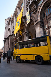 An arched neoclassical building with hanging banners, with a yellow vehicle parked in front