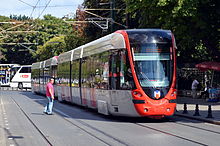 A red and silver electrified tram running through a street as a crossing pedestrian waits for it to pass
