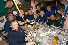 Thirteen astronauts seated around a table covered in open cans of food strapped down to the table. In the background a selection of equipment is visible, as well as the salmon-coloured walls of the Unity node.