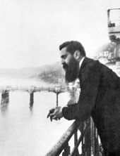 Um homem de barba longa em seus quarenta e poucos anos debruçado sobre uma grade com uma ponte no fundo.