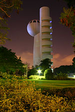 A night-time image of a gray windowless tower, with an egg-shaped windowed observation deck on top. Next to it is a low building, grass, and many trees and bushes.