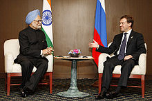 Two seated men converse. The first is dressed in Indian clothing and turban and sits before an Indian flag; the second is in a Western business suit and sits before a Russian flag.
