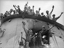 Feche acima do funil de um navio, que tem um grande buraco na lateral. Os marinheiros estão sorrindo e acenando para o fotógrafo do topo do funil e dentro do buraco.