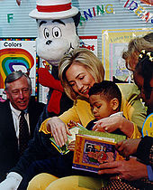 Mesma mulher lê um livro em uma sala de aula para um menino americano Africano em seu colo, como uma menina Africano americano e dois adultos olhar sobre