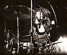 A black and white photograph of John Bonham wearing a headband and behind the cymbals of a drum kit