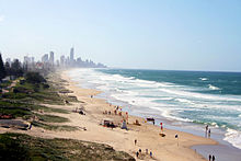 A beach sloping down from a grassy area on the left to the sea on the right, a city can be seen in the horizon