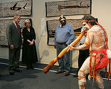 Aboriginal man performing on the Digeridoo indoors with 4 people watching, aboriginal paintings can be seen on the wall behind him