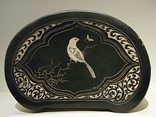 Um travesseiro em forma oval com lados planos. Ele é feito de pano preto com uma imagem de um pássaro branco sentado em um galho costurado para ele.