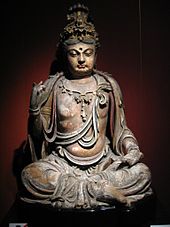 Uma escultura de madeira de um Buda pouco acima do peso, sentado em uma posição pernas cruzadas. Vestuário, incluindo uma camisa que cobre os ombros, mas deixa o peito exposto, e longas, calças largas, são esculpidas na estátua.