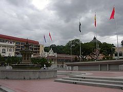 Uma fonte ornamentado na esquerda, com degraus que levam até uma parede com algumas das bandeiras do estado da Malásia sobre ele.