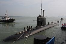 Um submarino Scorpène classe na doca, metade para fora da água. As pessoas no topo são amarração ele, e um barco pode ser visto no fundo