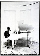 Uma foto preto e branco de Lennon sentada em um piano de cauda salão branco. Ele está usando fones de ouvido e uma camisa escura.