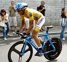 Um homem com roupas amarelas e um capacete azul, montando em uma bicicleta. No fundo alguns espectadores.
