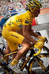 Um homem em roupas amarelas, com um capacete amarelo e handgloves amarelo, andar de bicicleta. Logo atrás dele está um outro ciclista, na roupa azul.