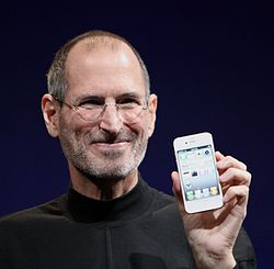 Retrato do ombro de alta do homem de sorriso em seus cinquenta anos vestindo uma camisa de gola alta preta com uma barba dias de idade, segurando um telefone de frente para o espectador em sua mão esquerda