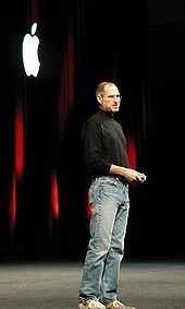 Retrato completo do homem sobre cinquenta usando jeans e uma camisa de gola alta preta, estando na frente de uma cortina escura com um logotipo da Apple branco