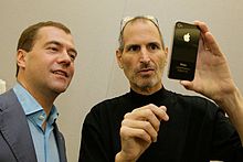 Shoulder-high portrait of two middle aged men, the one on left wearing a blue dress shirt and suitcoat, the one on right wearing a black turtleneck shirt and with his glasses pushed back onto his head and holding a phone facing them with an Apple logo visible on its back