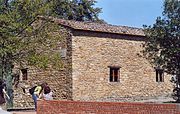 Foto de um edifício de pedra bruta com pequenas janelas, cercado por oliveiras.